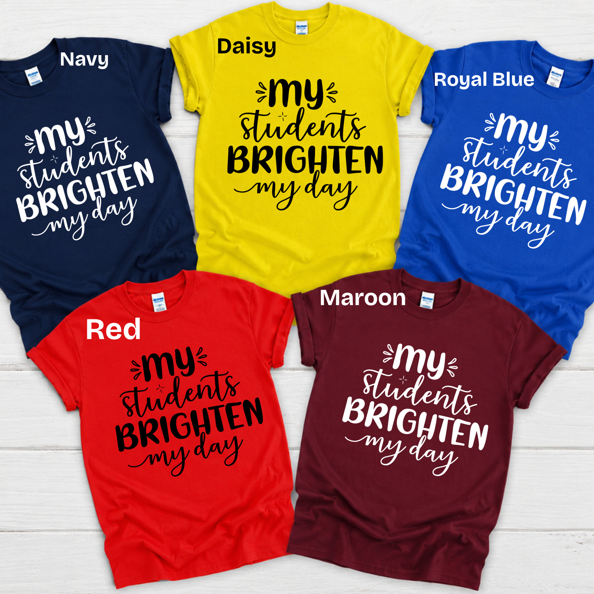 Six graphic t-shirts in different colors (Navy, Daisy yellow, Royal Blue, Red, Maroon) arranged in a circle on a white wood background. Each shirt features the text 'my students BRIGHTEN my day' in contrasting white or black lettering.