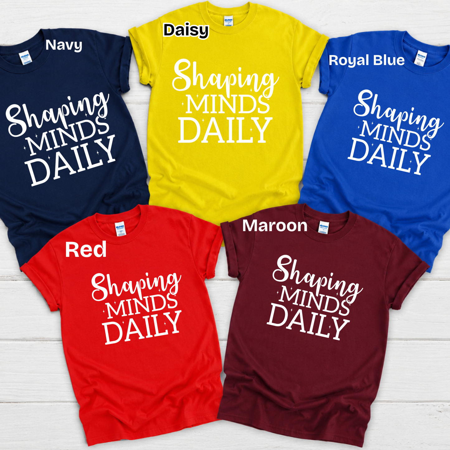 Six short-sleeve graphic T-shirts arranged in a 2x3 flat lay on a white wood background, each labeled with a color name: Navy, Daisy, Royal Blue, Red, Maroon, and a yellow shirt. The shirts all feature the phrase “TEACHING is my Super POWER” in white lettering.