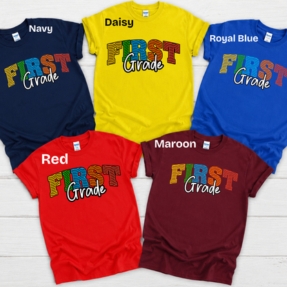 Five short-sleeve graphic T-shirts arranged in a fan layout on a white wood background, labeled Navy, Daisy, Royal Blue, Red, and Maroon. Each shirt has a colorful 'FIRST Grade' graphic printed across the chest.