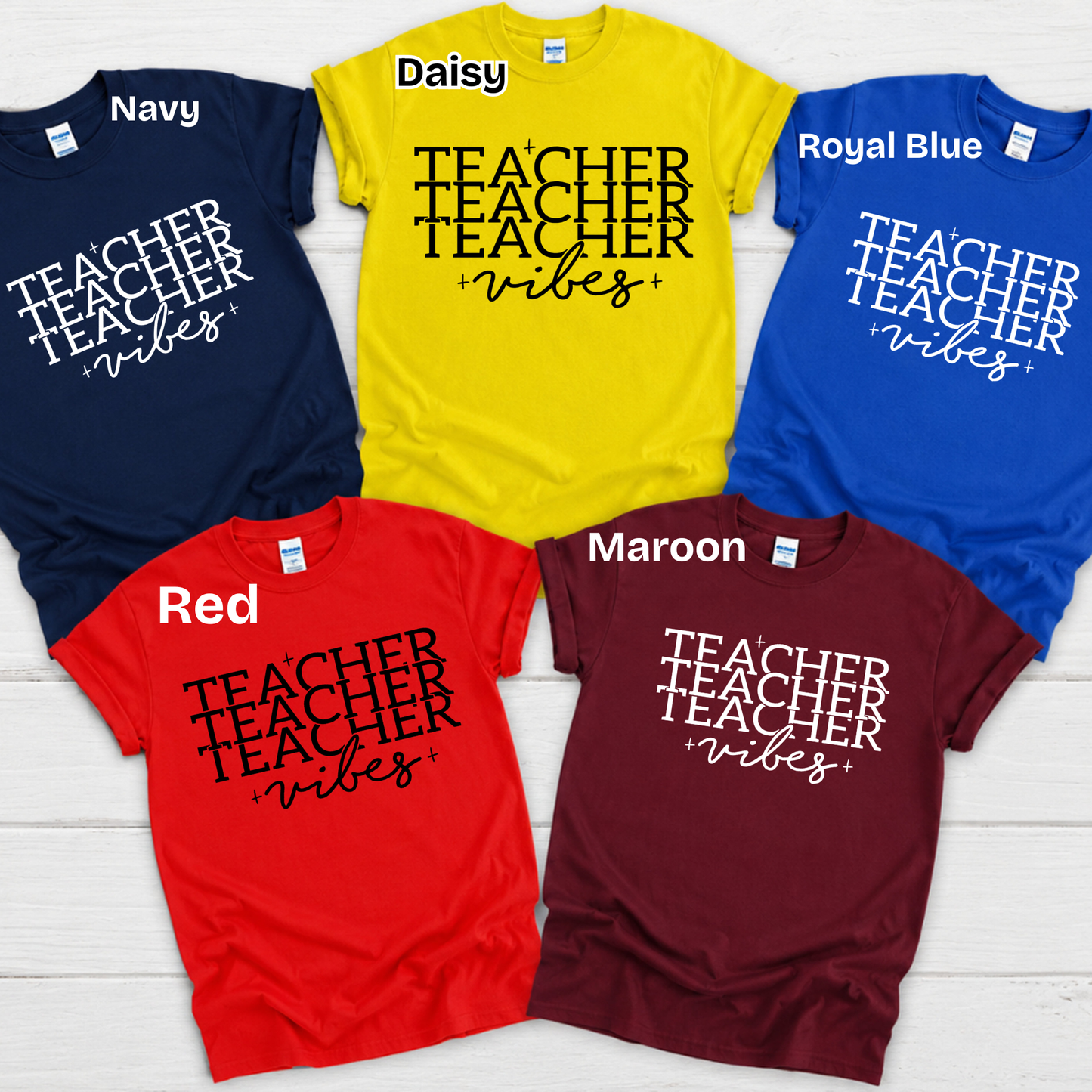Five graphic t-shirts in different colors are arranged in a flat-lay on a white wood background. The shirts are navy, yellow (labeled Daisy), royal blue, red, and maroon, all featuring the white printed text 'Shaping MINDS DAILY'.