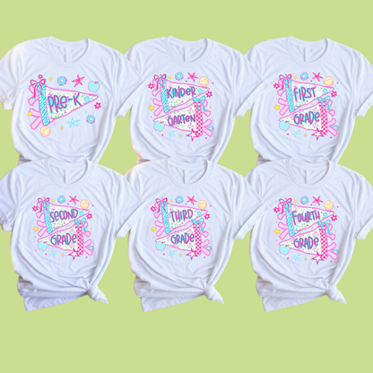 Six white short-sleeve T-shirts arranged in two rows on a pale yellow-green background. Each shirt has a colorful graphic print with school-grade-themed text and playful doodles including stars, hearts, and ribbon motifs.
