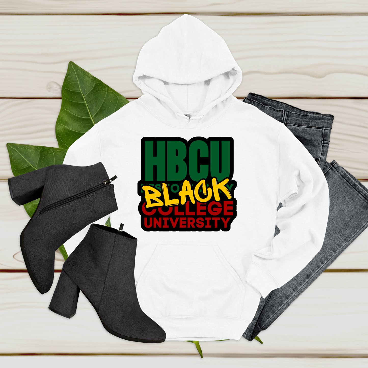 White hoodie with 'HBCU Black College University' text, black boots, and jeans on a wooden surface.