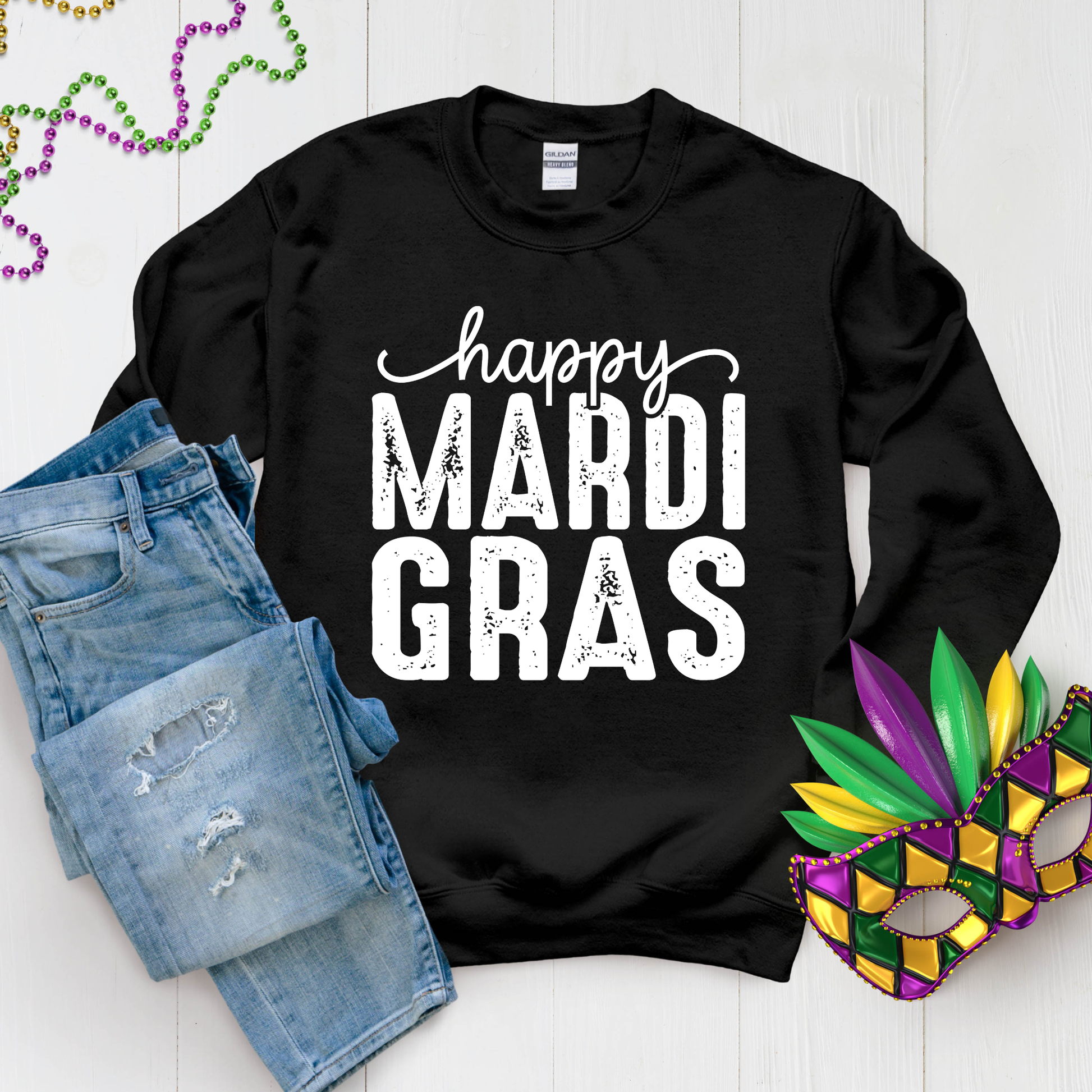 Black sweatshirt with 'happy Mardi Gras' text, denim shorts, and a Mardi Gras mask on a light wooden surface.