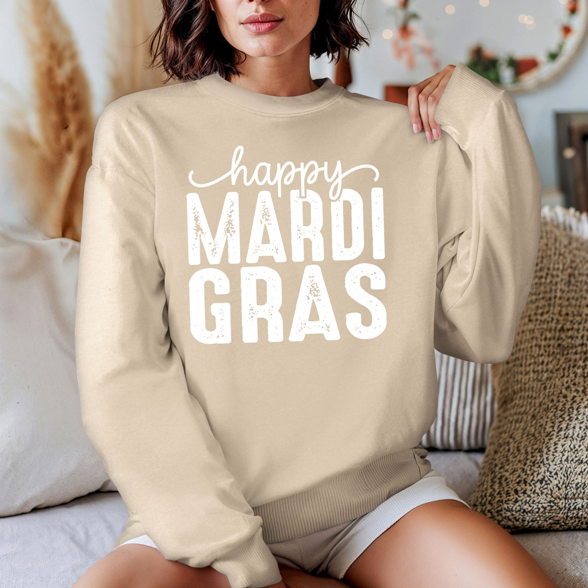 Person wearing a beige sweatshirt with 'happy MARDI GRAS' text in a cozy indoor setting.