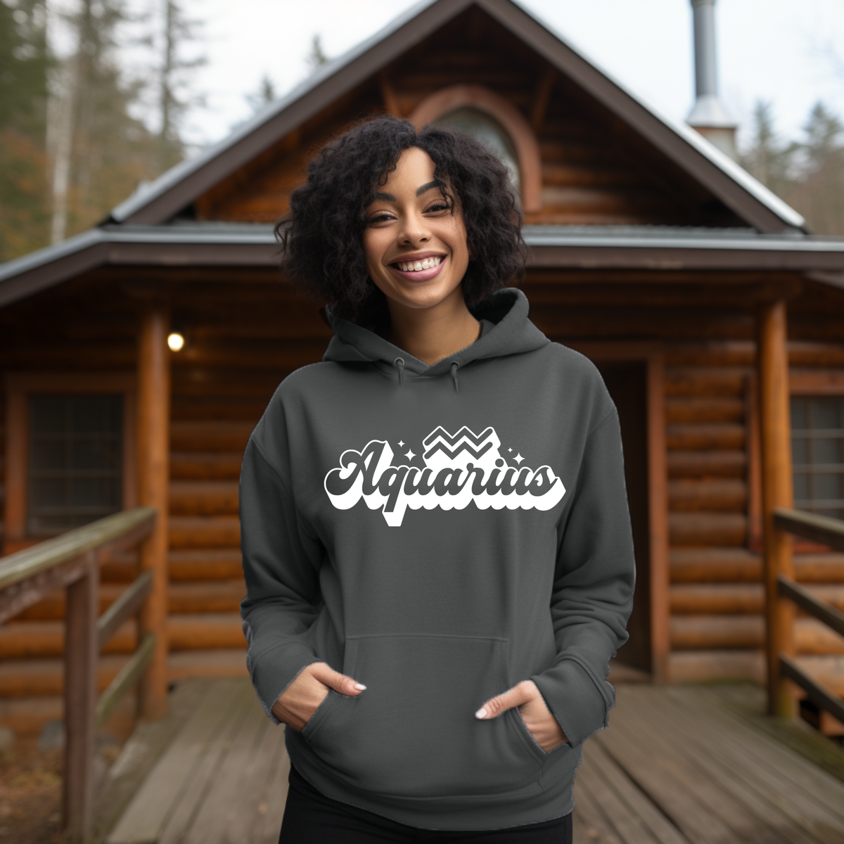 A smiling woman with curly black hair stands on a wooden porch in front of a log cabin, wearing a dark charcoal gray hoodie with 'Aquarius' printed in white script across the chest.