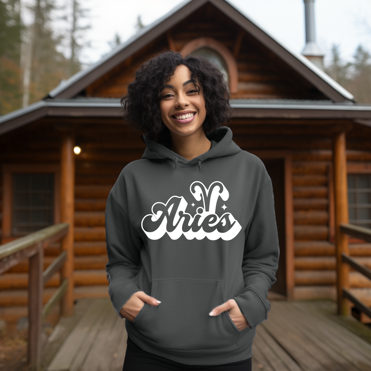 A woman wearing a dark gray hoodie with a white 'Aries' graphic stands smiling on a wooden porch in front of a log cabin.
