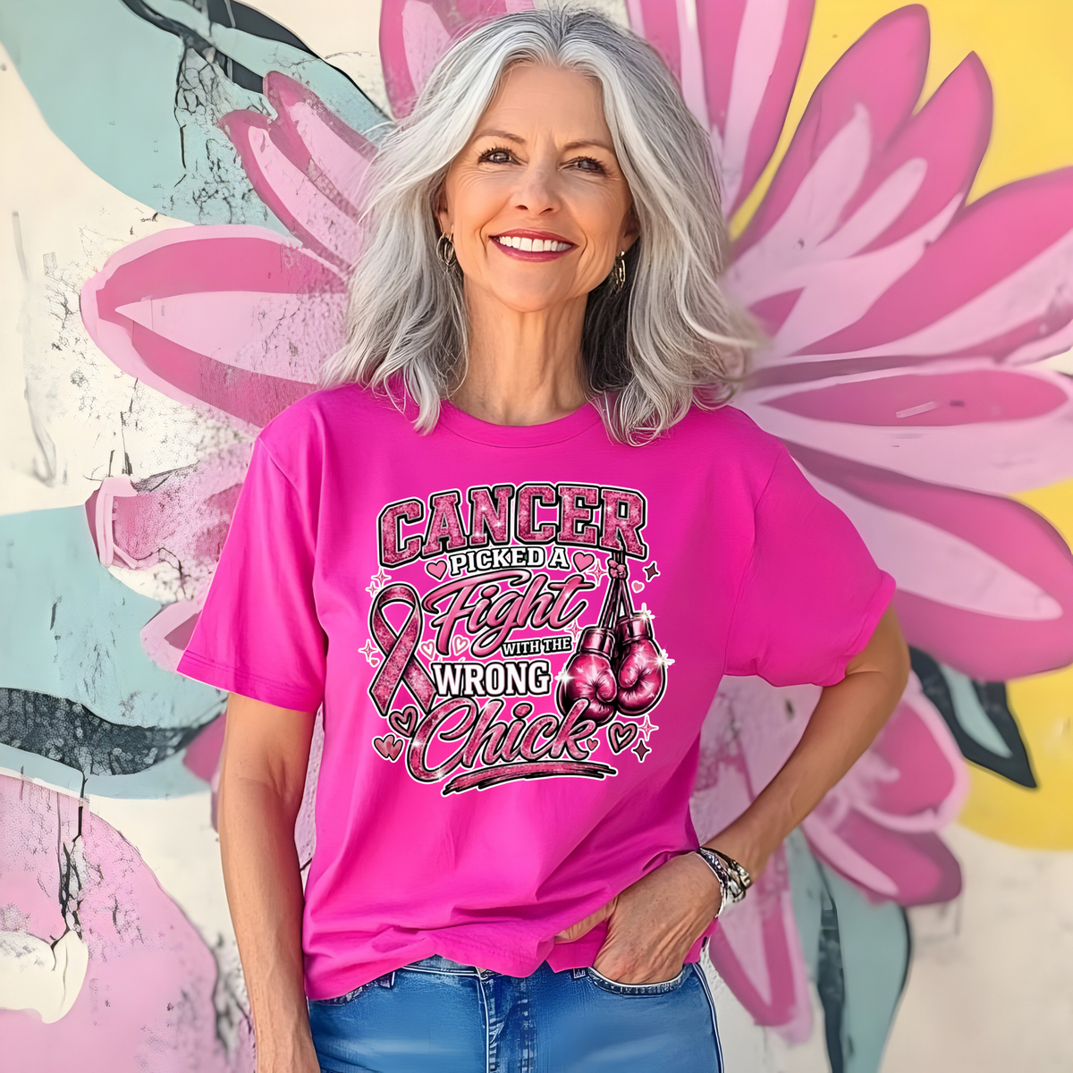 A smiling woman with gray hair stands in front of a colorful floral wall mural, wearing a bright pink graphic T-shirt with the text 'Cancer Picked a Fight and I'm Not Wrong Chick' and a ribbon/cherry design, paired with blue jeans.