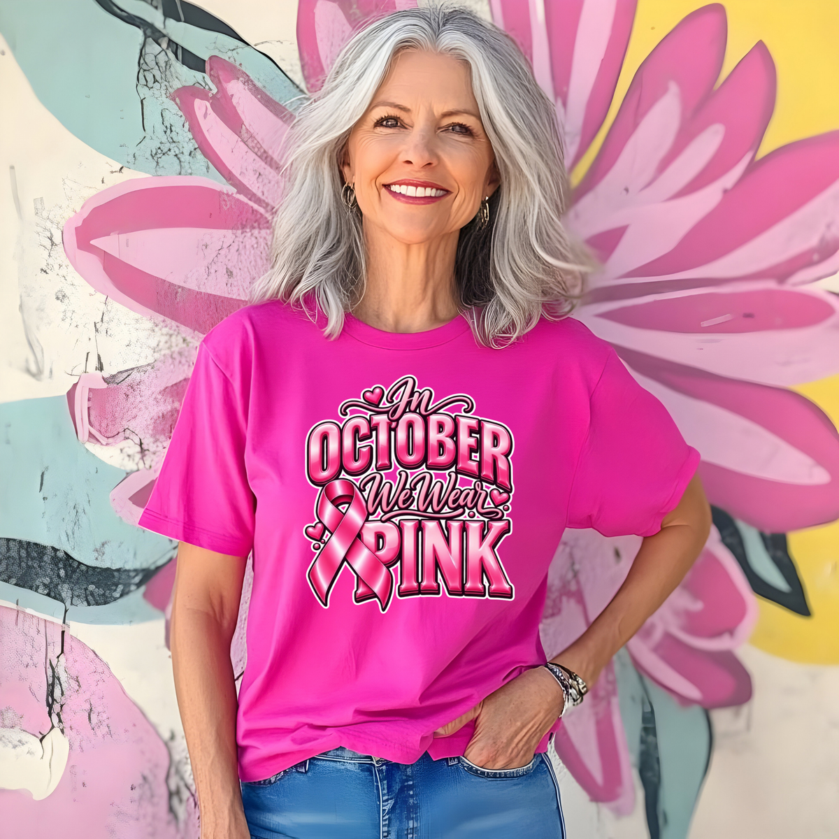 A smiling woman with gray hair wears a bright pink graphic T-shirt featuring a pink breast cancer ribbon, pink boxing gloves, and the words 'Breast Cancer WARRIOR' in black and white. She is standing in front of a colorful floral mural.