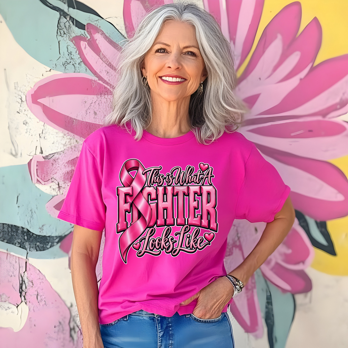 A smiling woman with gray hair wears a bright pink graphic T-shirt featuring a pink breast cancer ribbon, pink boxing gloves, and the words 'Breast Cancer WARRIOR' in black and white. She is standing in front of a colorful floral mural.