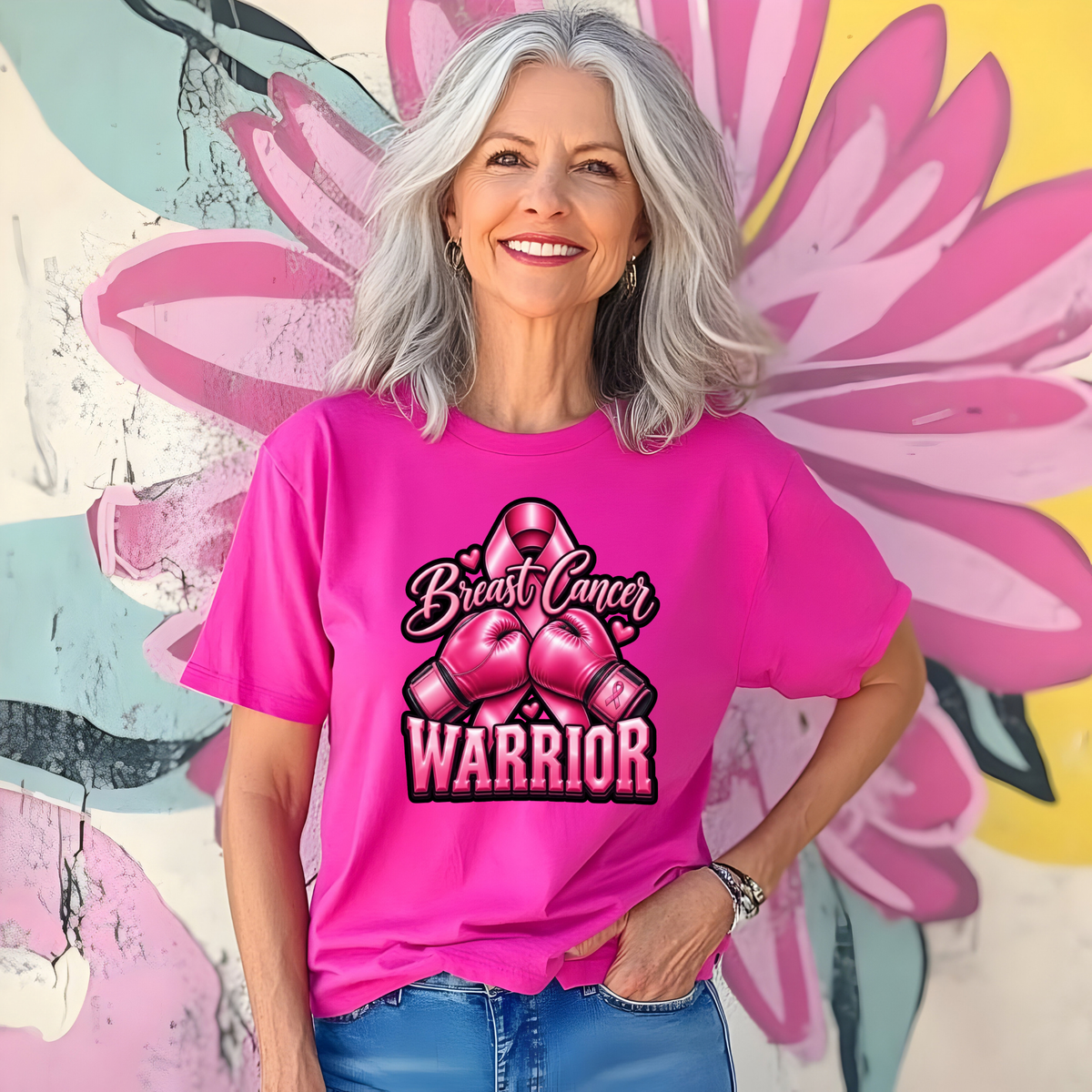 A smiling woman with gray hair wears a bright pink graphic T-shirt featuring a pink breast cancer ribbon, pink boxing gloves, and the words 'Breast Cancer WARRIOR' in black and white. She is standing in front of a colorful floral mural.