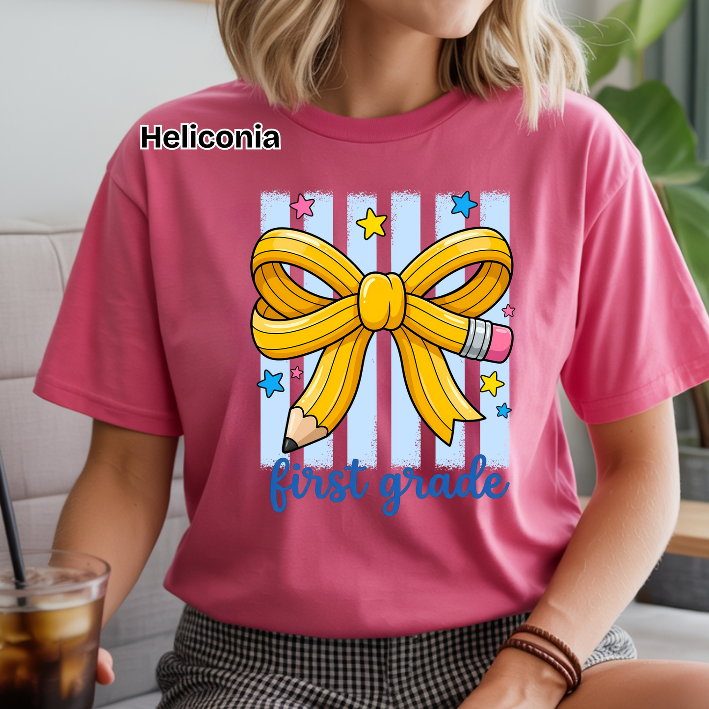 A woman wearing a pink graphic t-shirt with a yellow ribbon and pencil design that says 'Heliconia first grade'.