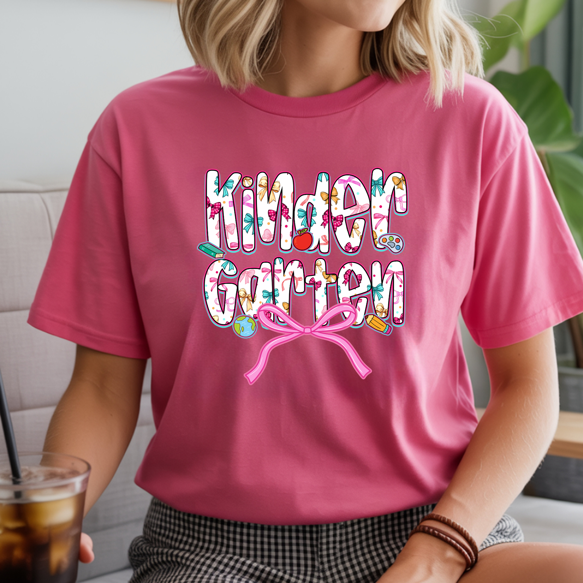 A woman wearing a pink short-sleeve graphic T-shirt with the words 'Kinder Garten' in a colorful floral pattern and a pink bow underneath, shown in a casual indoor setting.