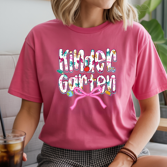 A woman wearing a pink short-sleeve graphic T-shirt with the words 'Kinder Garten' in a colorful floral pattern and a pink bow underneath, shown in a casual indoor setting.