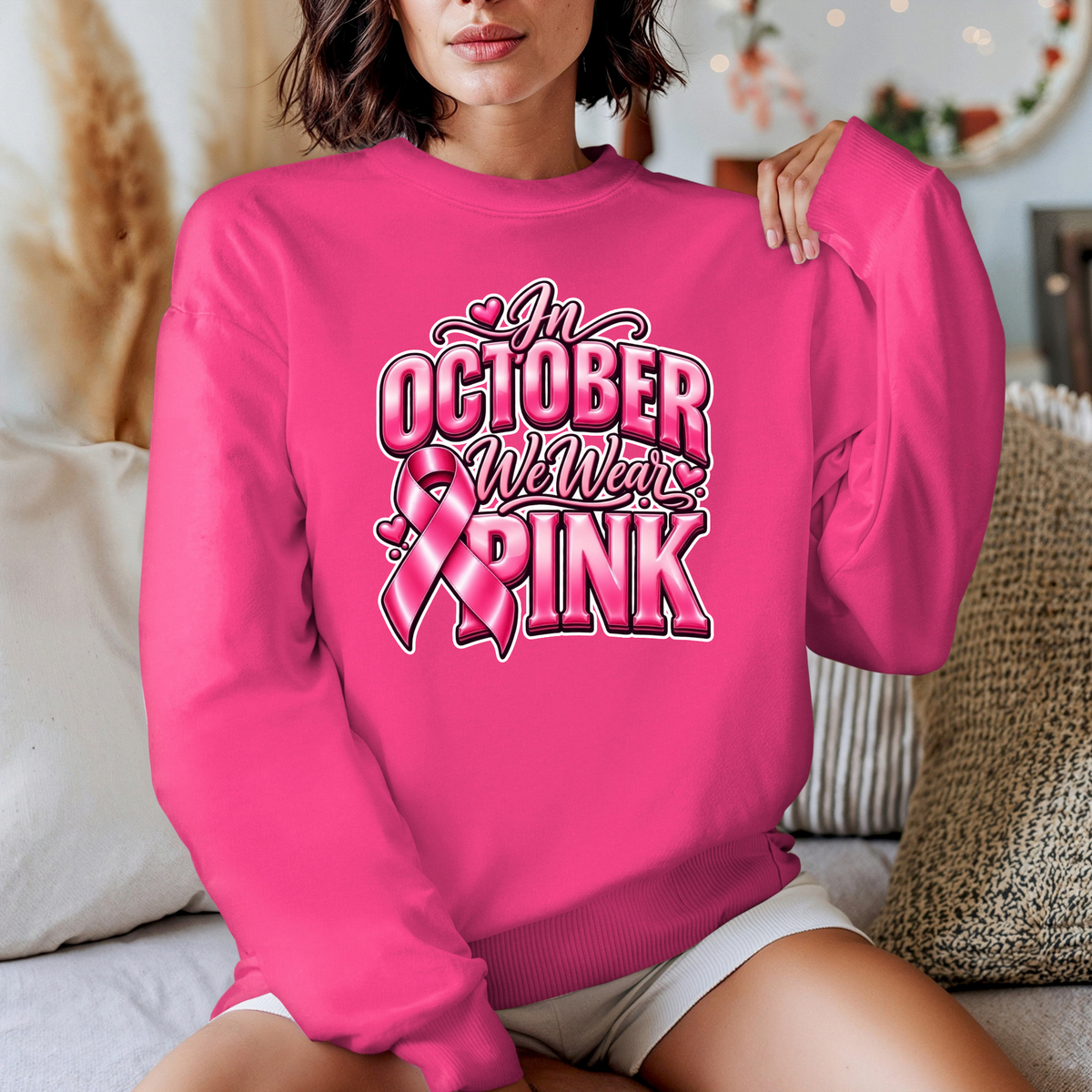 A woman wearing a bright pink crewneck sweatshirt with a graphic that reads 'Breast Cancer Warrior' in script and block lettering, featuring pink boxing gloves and a pink ribbon. The photo is taken in a cozy indoor setting with pillows in the background.
