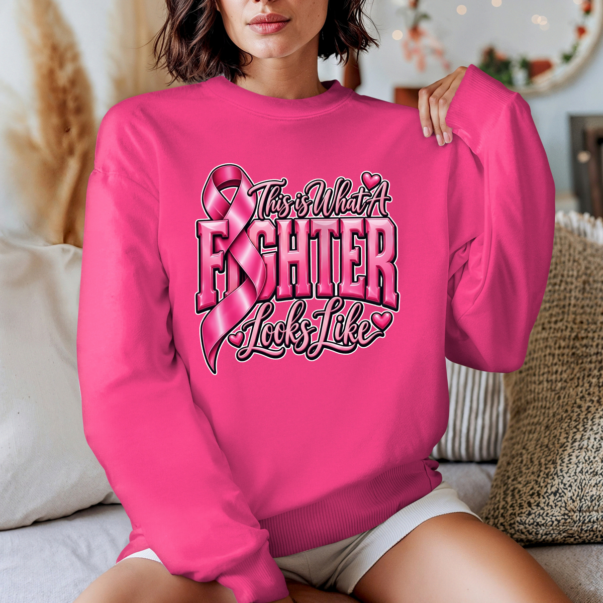 A woman wearing a bright pink crewneck sweatshirt with a graphic that reads 'Breast Cancer Warrior' in script and block lettering, featuring pink boxing gloves and a pink ribbon. The photo is taken in a cozy indoor setting with pillows in the background.