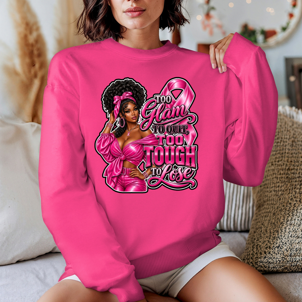 A woman wearing a bright pink crewneck sweatshirt with a graphic that reads 'Breast Cancer WARRIOR' in script and block lettering, featuring pink boxing gloves and a pink ribbon. The photo is taken in a cozy indoor setting with pillows in the background.