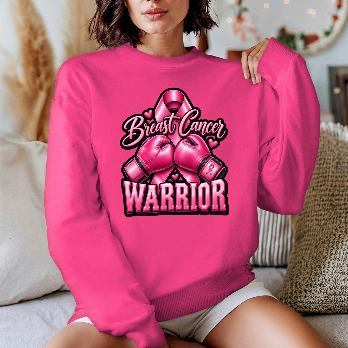 A woman wearing a bright pink crewneck sweatshirt with a graphic that reads 'Breast Cancer Warrior' in script and block lettering, featuring pink boxing gloves and a pink ribbon. The photo is taken in a cozy indoor setting with pillows in the background.