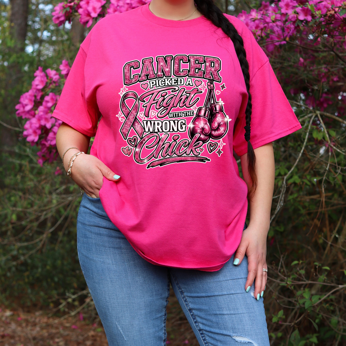 A woman wearing a bright pink graphic t-shirt with the text 'Cancer Picked a Fight with the Wrong Chick' and a ribbon/cherries design, paired with blue jeans, standing outdoors in front of pink flowers and green foliage.