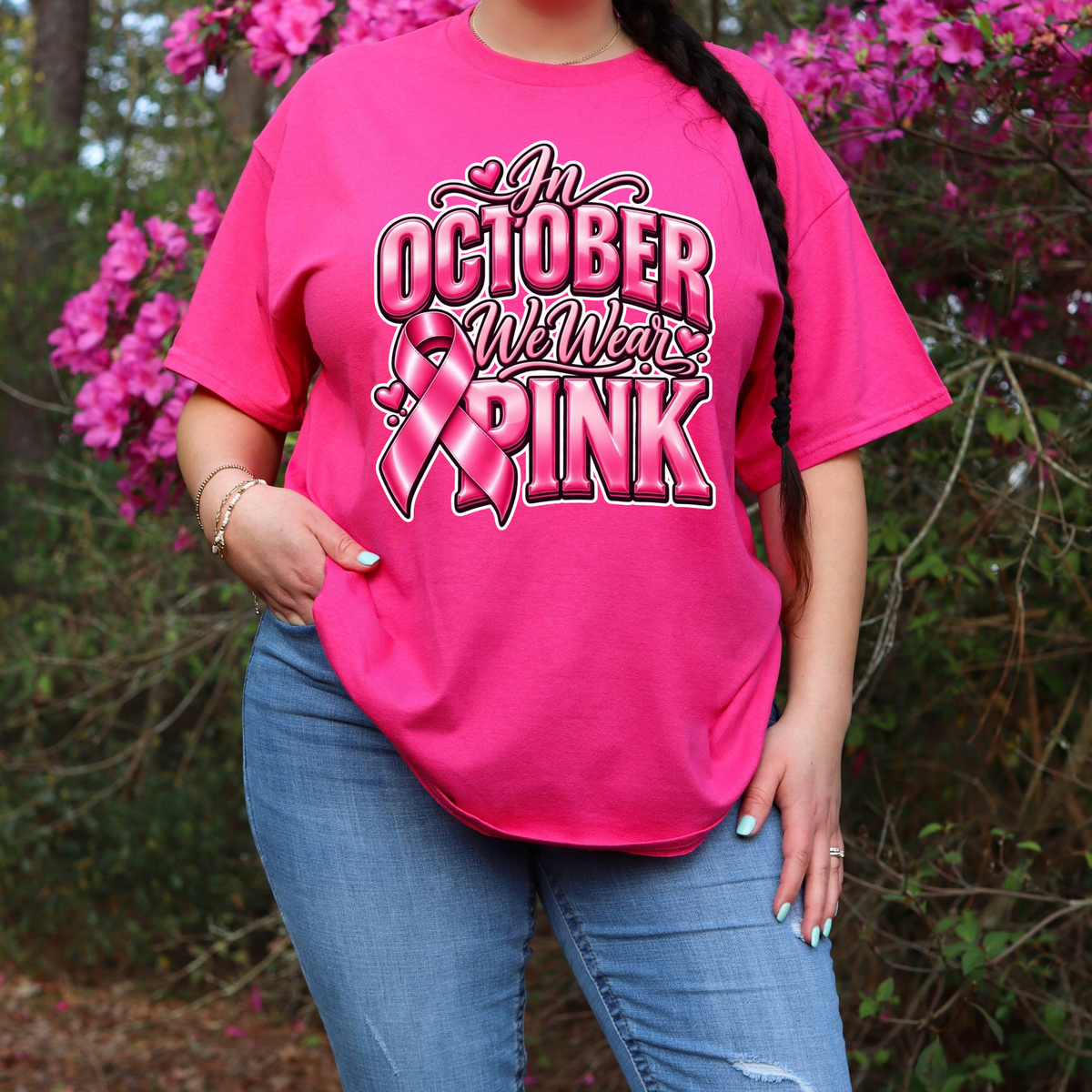 A woman wearing a bright pink graphic t-shirt with a 'Breast Cancer Warrior' boxing gloves and ribbon design, paired with blue jeans, standing outdoors in front of pink flowering bushes.