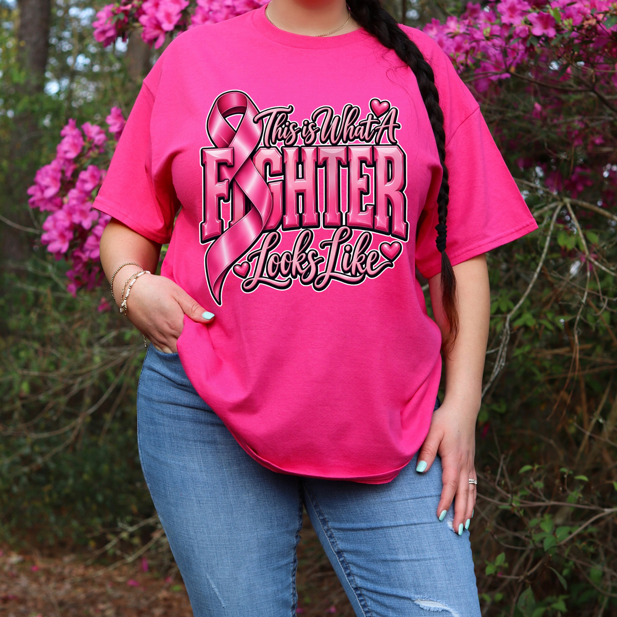 A woman wearing a bright pink graphic t-shirt with a 'Breast Cancer Warrior' boxing gloves and ribbon design, paired with blue jeans, standing outdoors in front of pink flowering bushes.