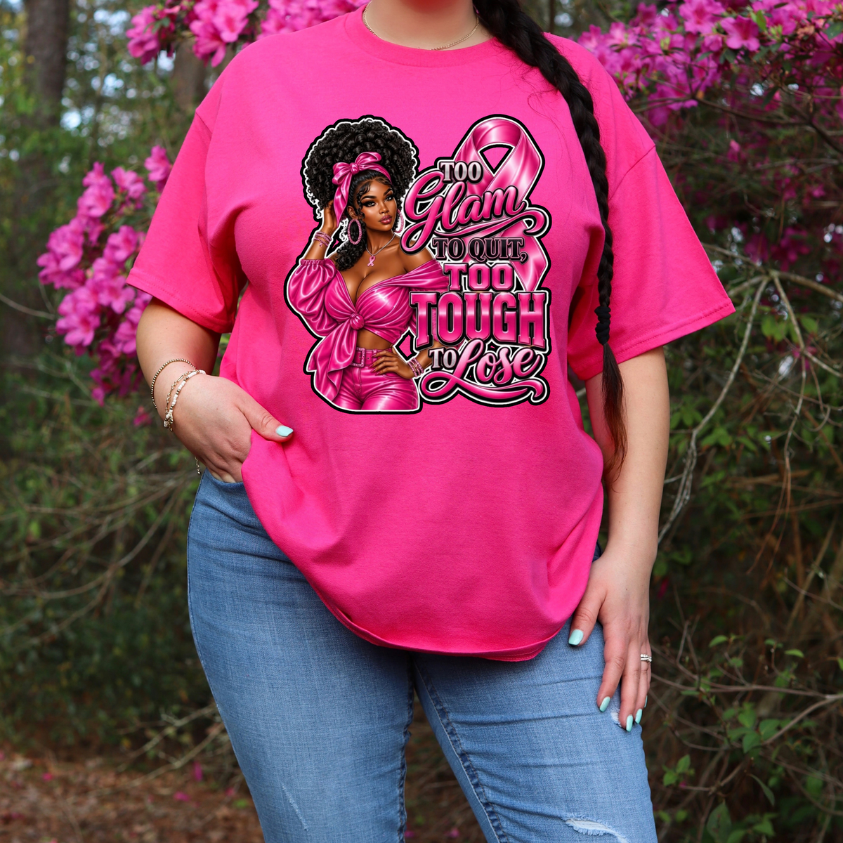 A woman wearing a bright pink graphic t-shirt with a 'Breast Cancer Warrior' boxing gloves and ribbon design, paired with blue jeans, standing outdoors in front of pink flowering bushes.