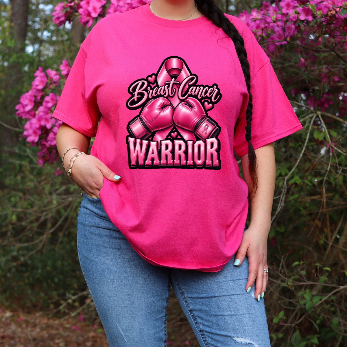 A woman wearing a bright pink graphic t-shirt with a 'Breast Cancer Warrior' boxing gloves and ribbon design, paired with blue jeans, standing outdoors in front of pink flowering bushes.