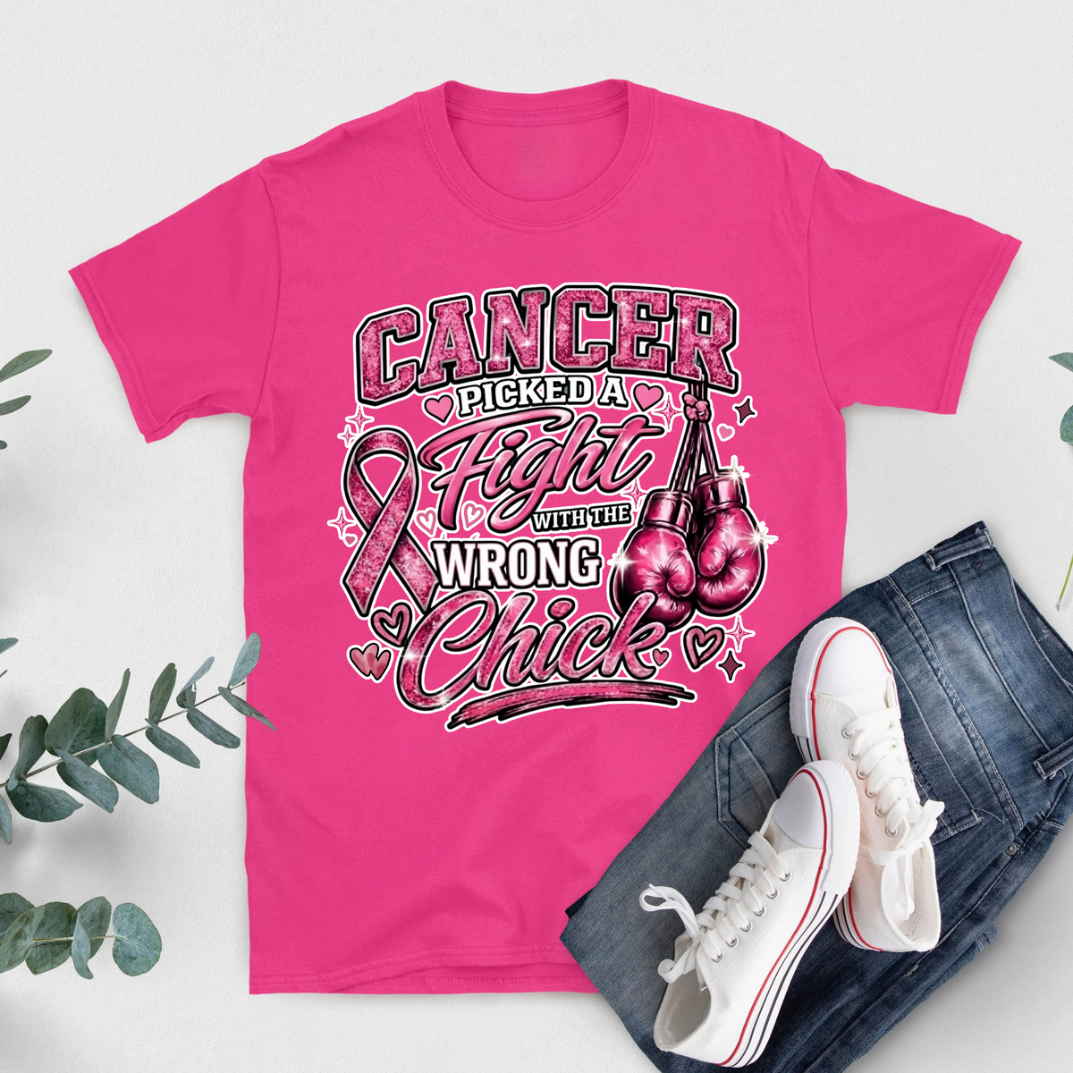 A bright pink graphic T-shirt laid flat on a light gray background, styled with blue jeans and white sneakers. The shirt features a large printed design with three women, a pink ribbon, and the text 'HER Fight IS Our Fight'.
