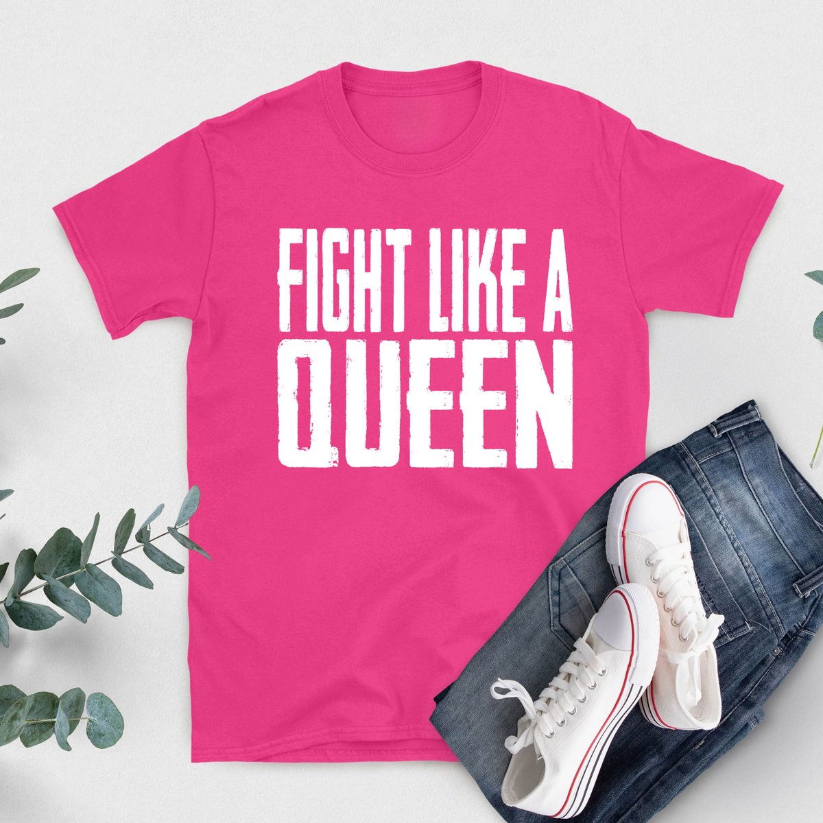 A bright pink short-sleeve T-shirt with bold white text reading 'FIGHT LIKE A QUEEN' laid flat on a light background, styled with blue jeans and white sneakers with red stripes, plus green leaves around the edges.