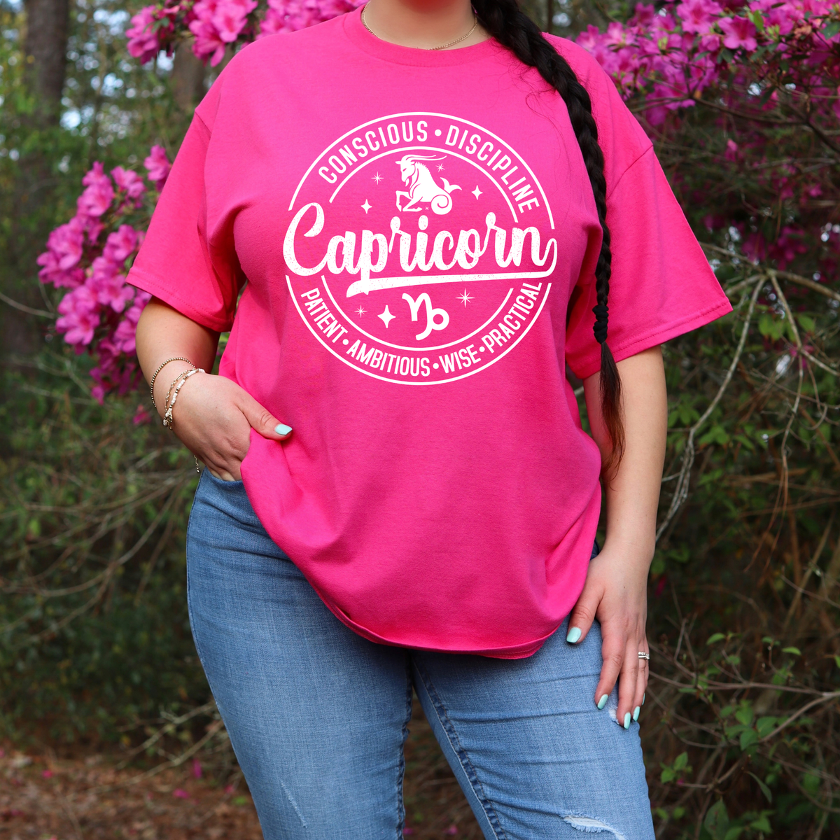A woman wearing a bright pink graphic T-shirt with a black circular design that reads 'Libra' and additional text, paired with blue jeans, standing outdoors in front of pink flowers and green foliage.