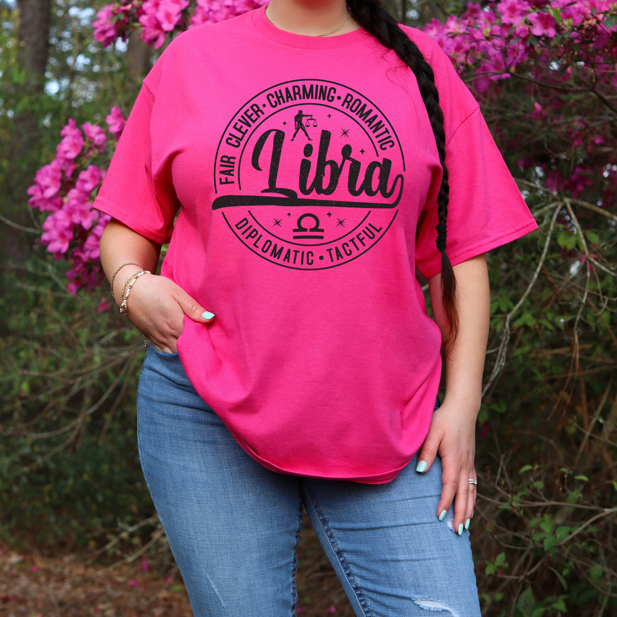 A woman wearing a bright pink graphic T-shirt with a black circular design that reads 'Libra' and additional text, paired with blue jeans, standing outdoors in front of pink flowers and green foliage.