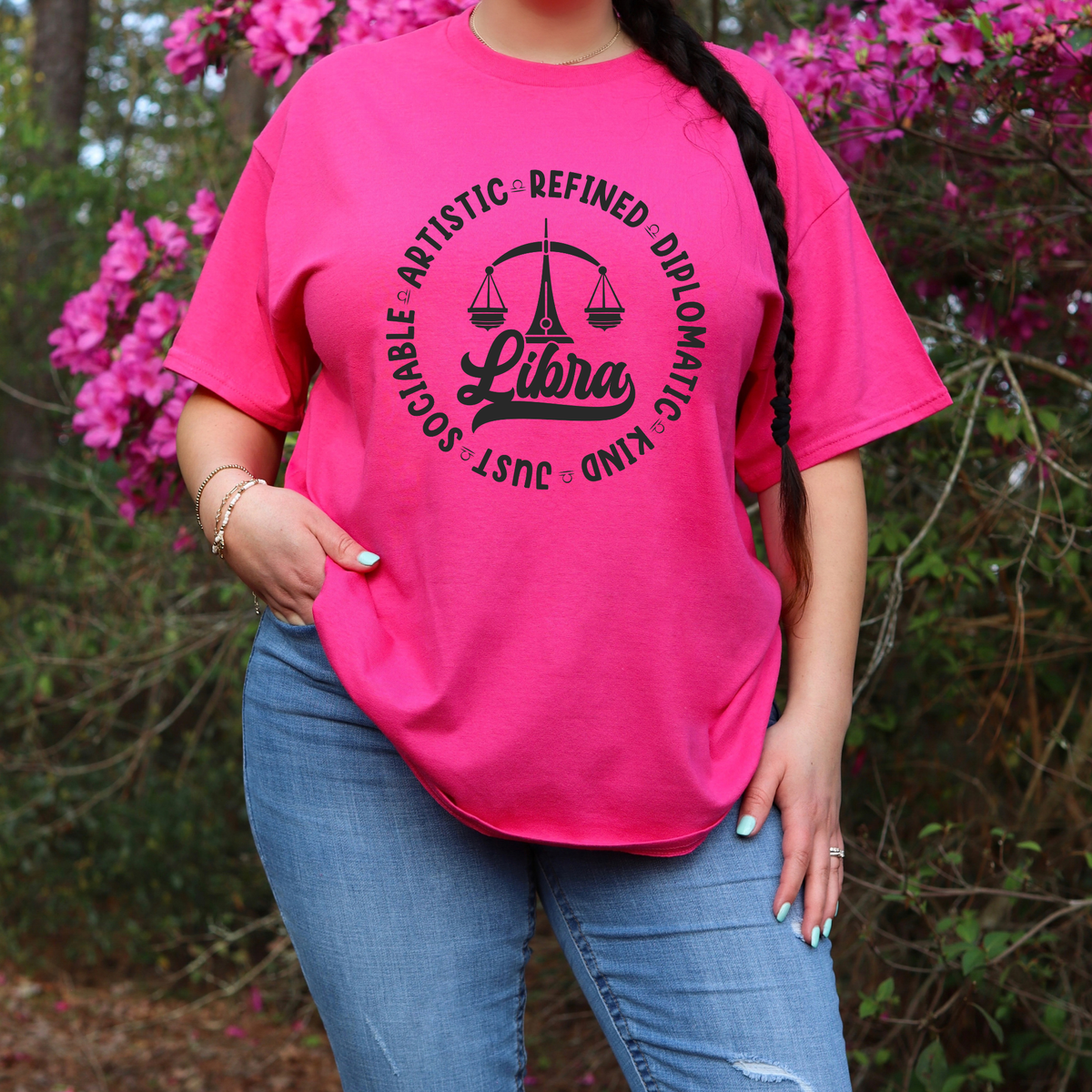 A woman wearing a bright pink t-shirt with a black Libra graphic print, paired with blue jeans, standing outdoors in front of pink flowers and green foliage.