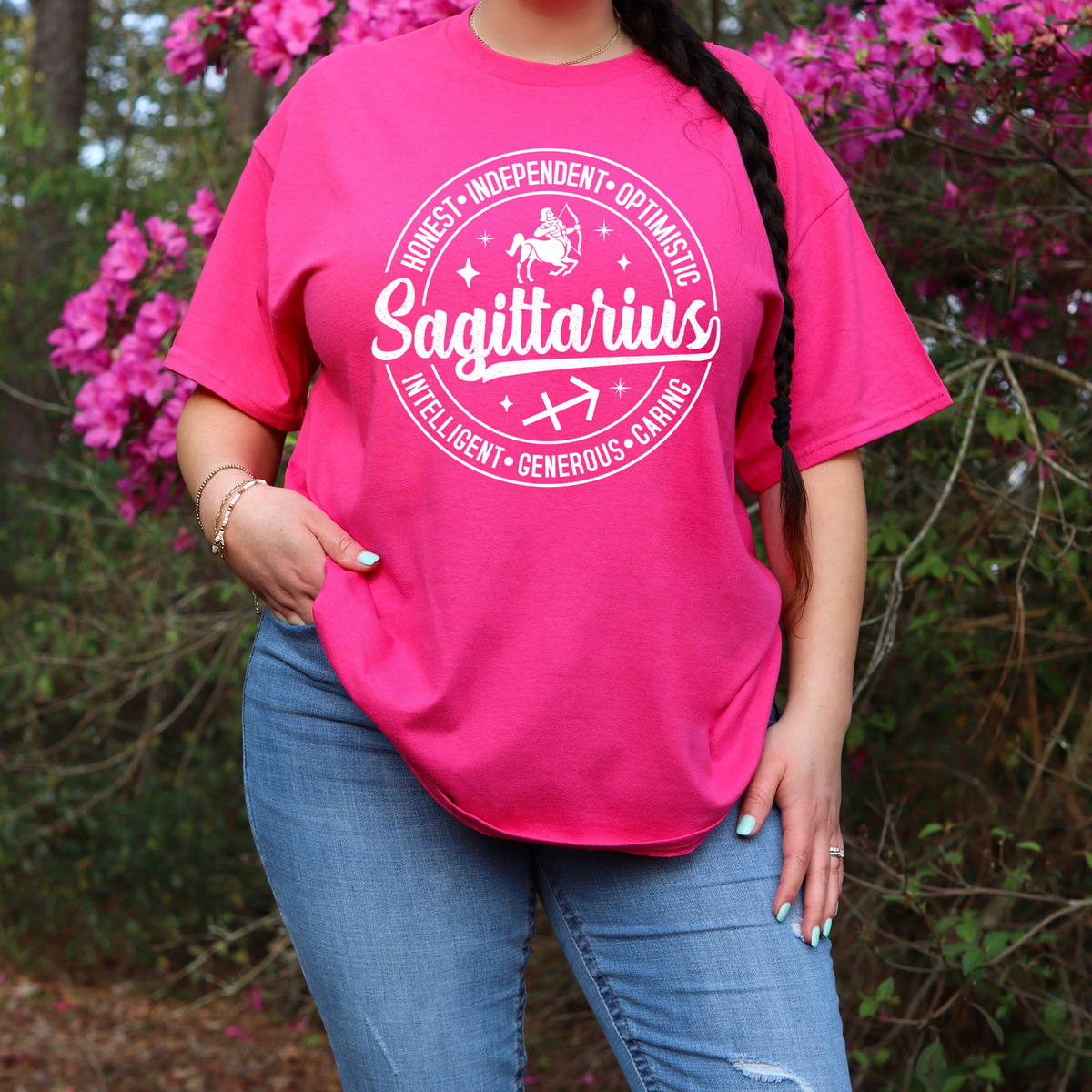 A woman wearing a bright pink graphic T-shirt with a black circular design that reads 'Libra' and additional text, paired with blue jeans, standing outdoors in front of pink flowers and green foliage.
