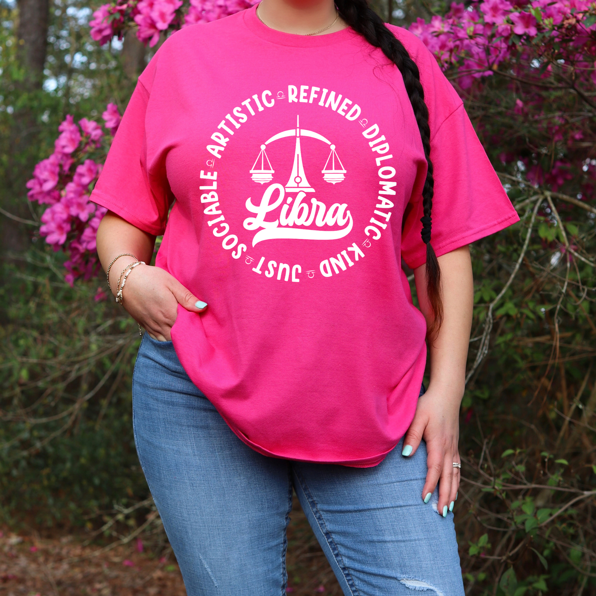 Woman wearing a white crewneck t-shirt featuring a black circular Libra zodiac graphic with a scales illustration, the word "Libra," and trait keywords like artistic, refined, diplomatic, kind, just, and sociable.