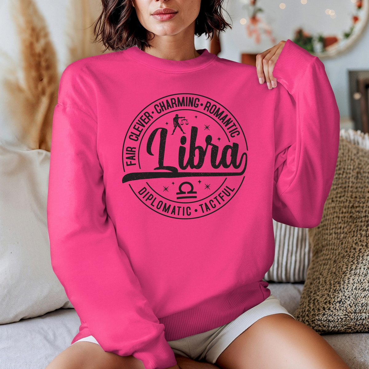 A woman wearing a bright pink crewneck sweatshirt with a black graphic print that reads 'Libra' with descriptive adjectives around it, seated in a cozy indoor setting.