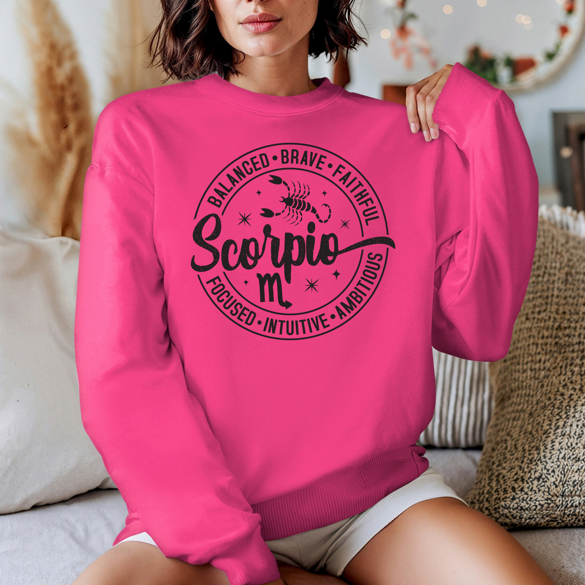 A woman wearing a bright pink crewneck sweatshirt with a black graphic print that reads 'Libra' with descriptive adjectives around it, seated in a cozy indoor setting.
