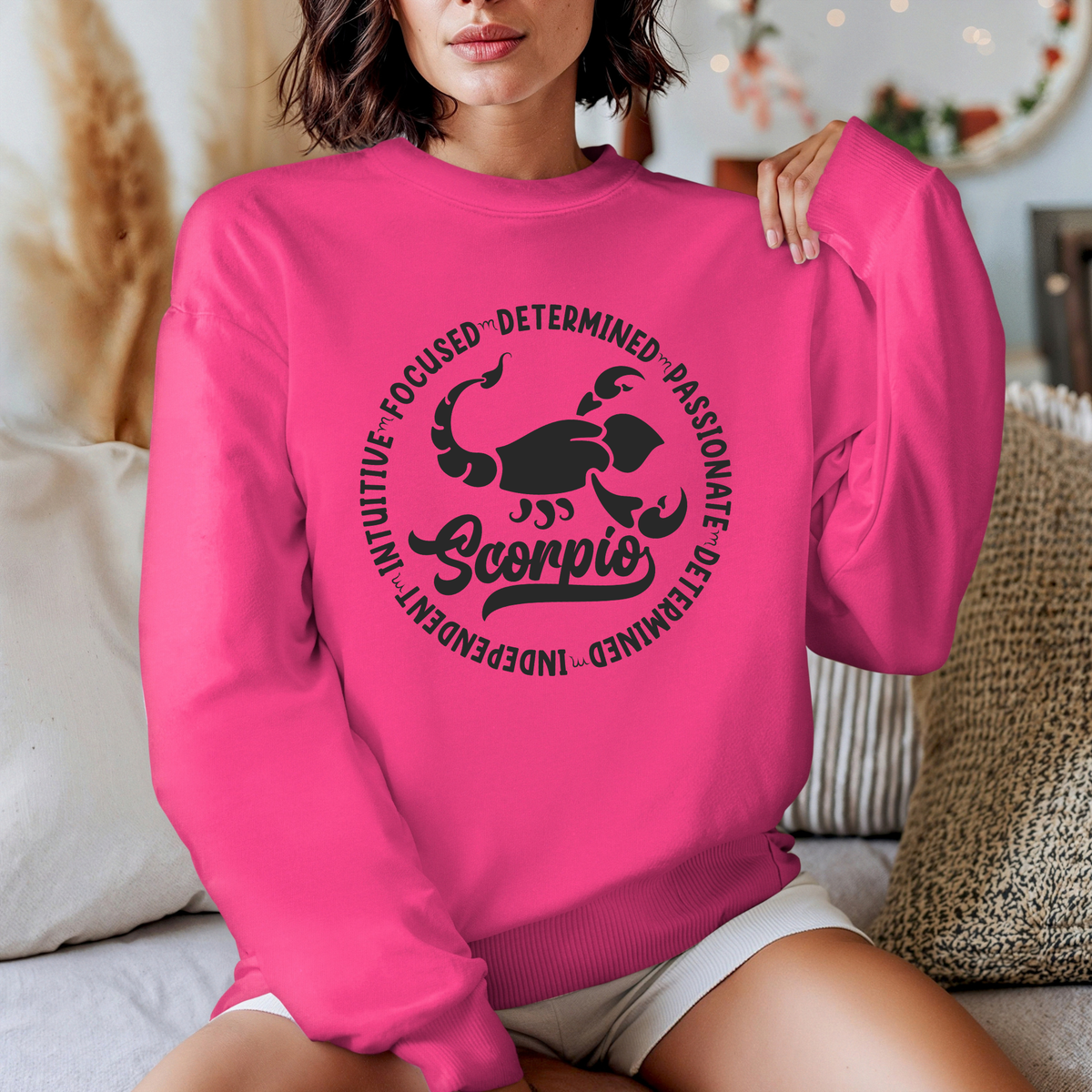 Woman wearing a black crewneck sweatshirt featuring a white circular Scorpio zodiac graphic with a scorpion illustration, the word "Scorpio," and trait keywords like intuitive, focused, determined, passionate, and independent.