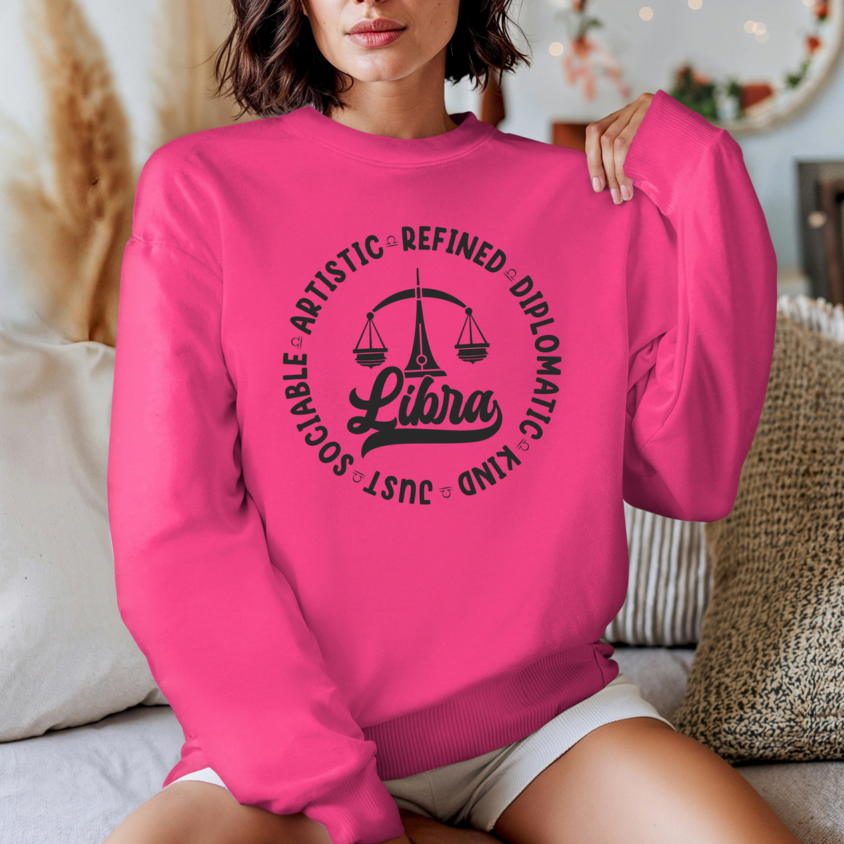 Woman wearing a white crewneck t-shirt featuring a black circular Libra zodiac graphic with a scales illustration, the word "Libra," and trait keywords like artistic, refined, diplomatic, kind, just, and sociable.