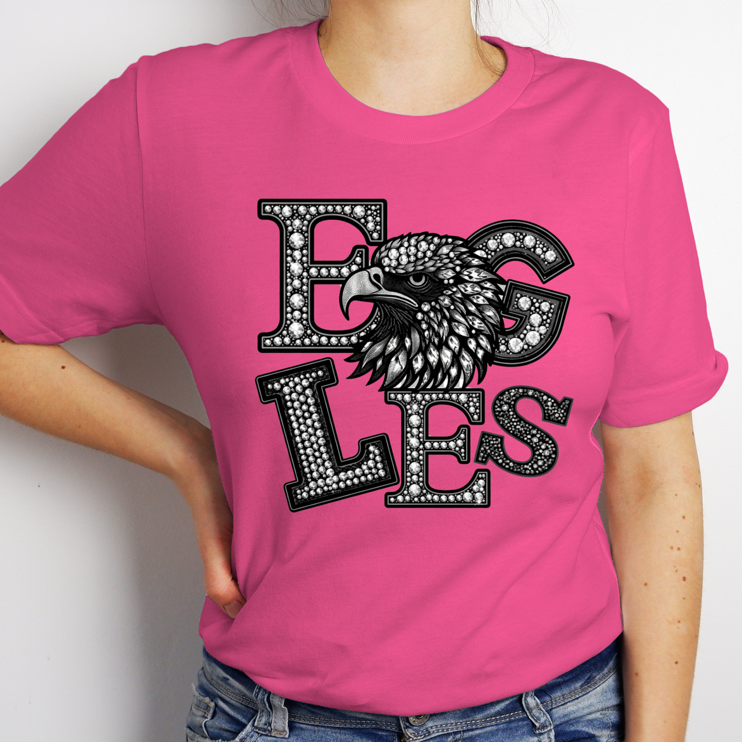 A woman wearing a bright pink short-sleeve t-shirt with a black and white graphic that reads 'EAGLES' in a textured, gemstone-like font, featuring an eagle head design. The model is also wearing blue jeans.