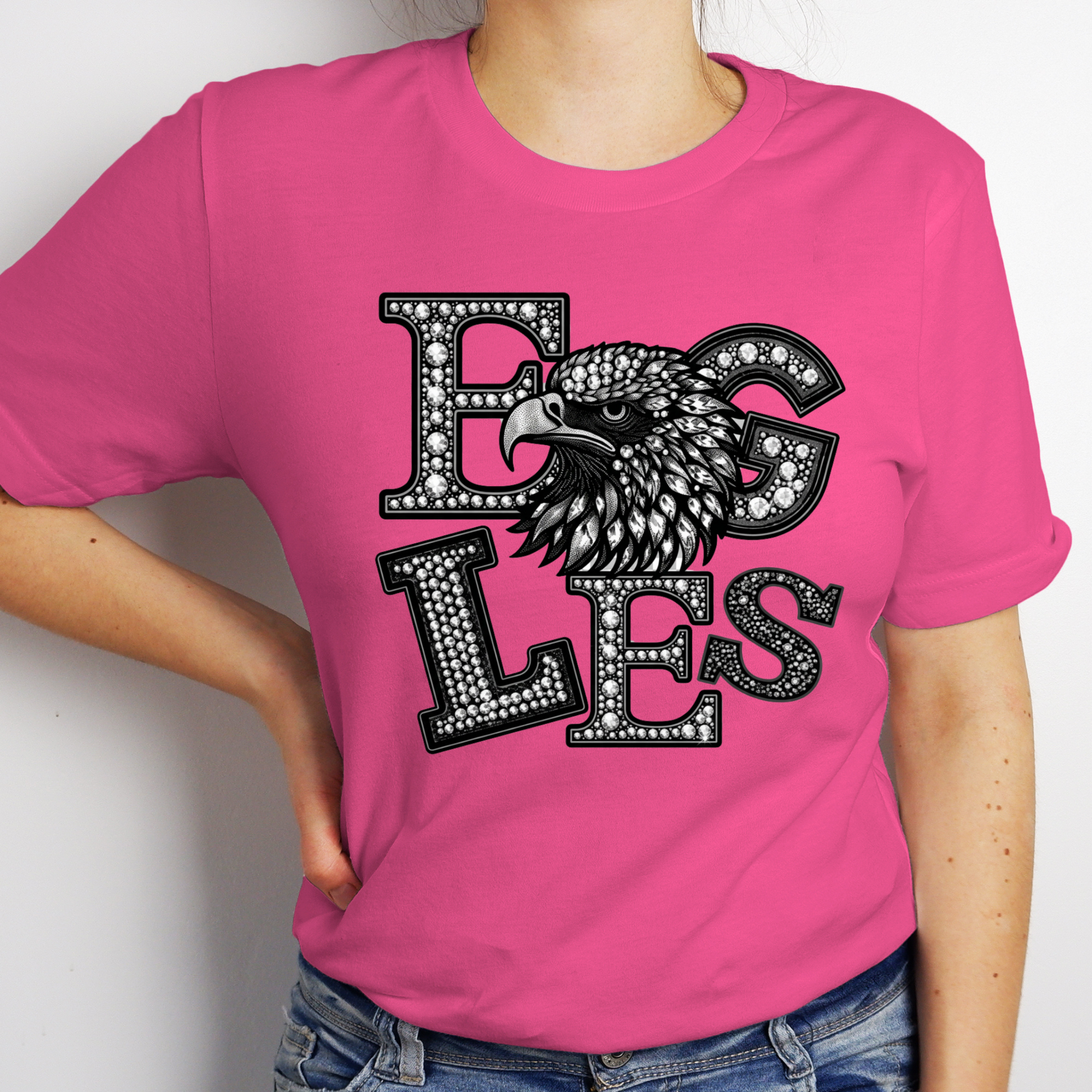 A woman wearing a bright pink short-sleeve t-shirt with a black and white graphic that reads 'EAGLES' in a textured, gemstone-like font, featuring an eagle head design. The model is also wearing blue jeans.