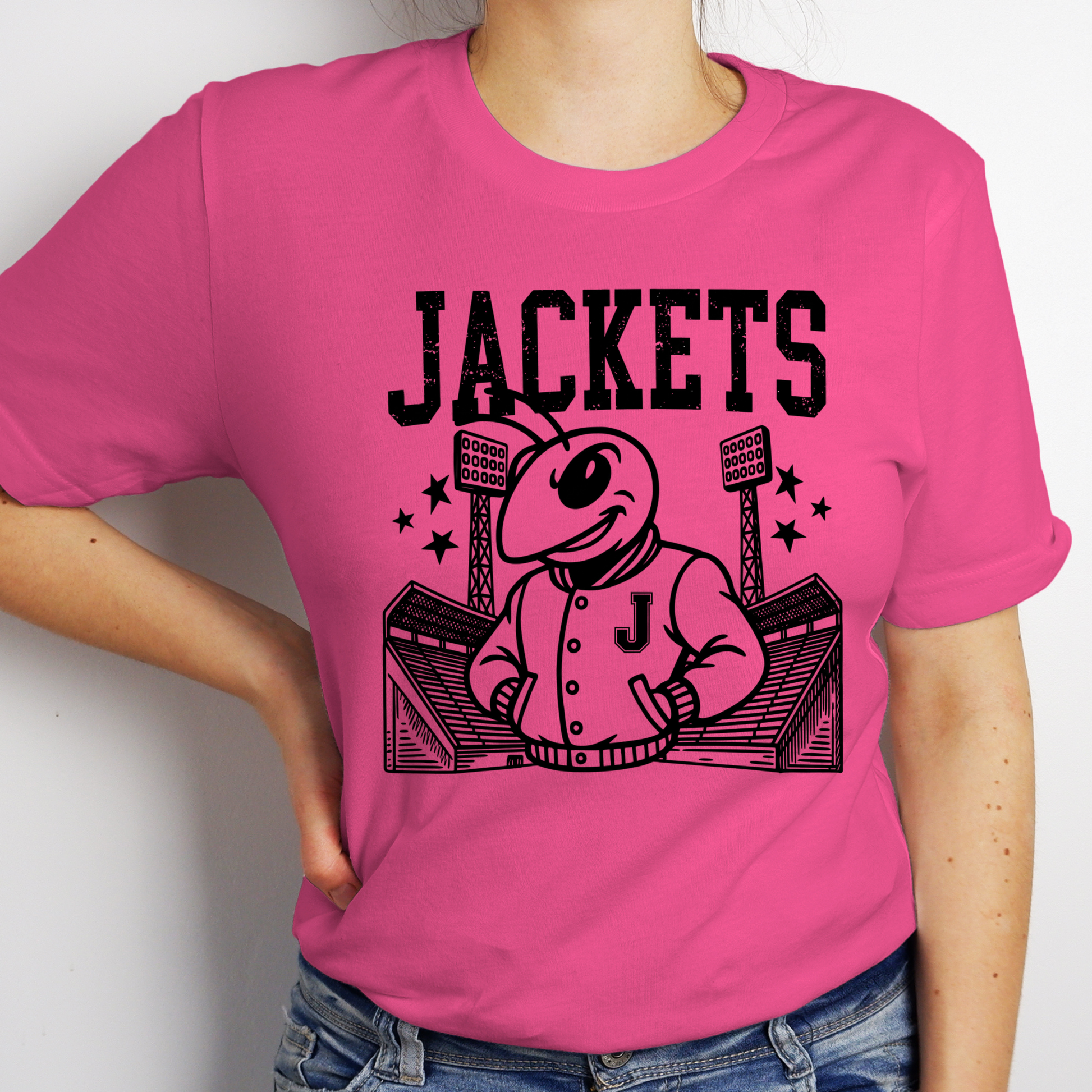 A woman wearing a bright pink short-sleeve t-shirt with a black graphic of a mascot wearing a jacket with the letter 'J' and the word 'JACKETS' above it, paired with blue jeans.