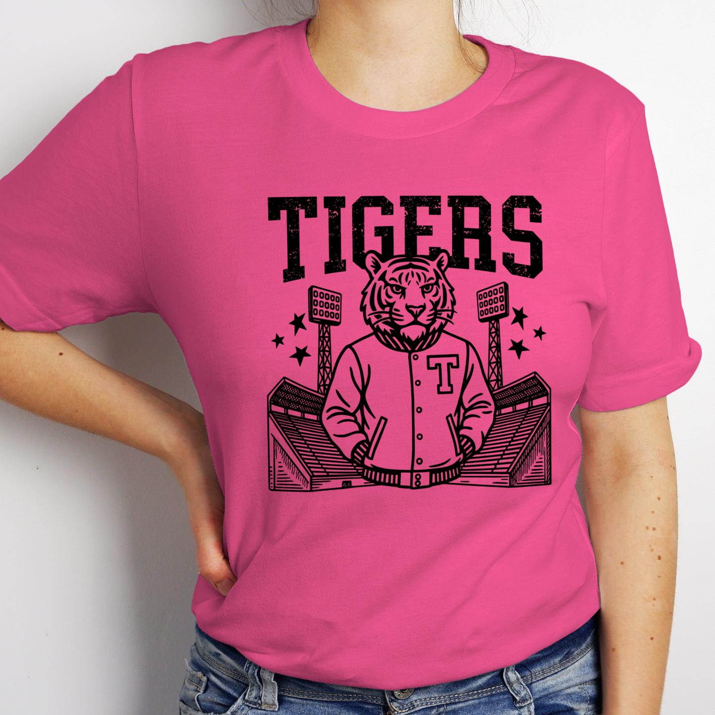 A person wearing a bright pink short-sleeve T-shirt with a large black graphic on the front showing the word "TIGERS" above an illustrated tiger in a varsity jacket standing in stadium seating. The photo is cropped from the neck to the hips against a plain light background.