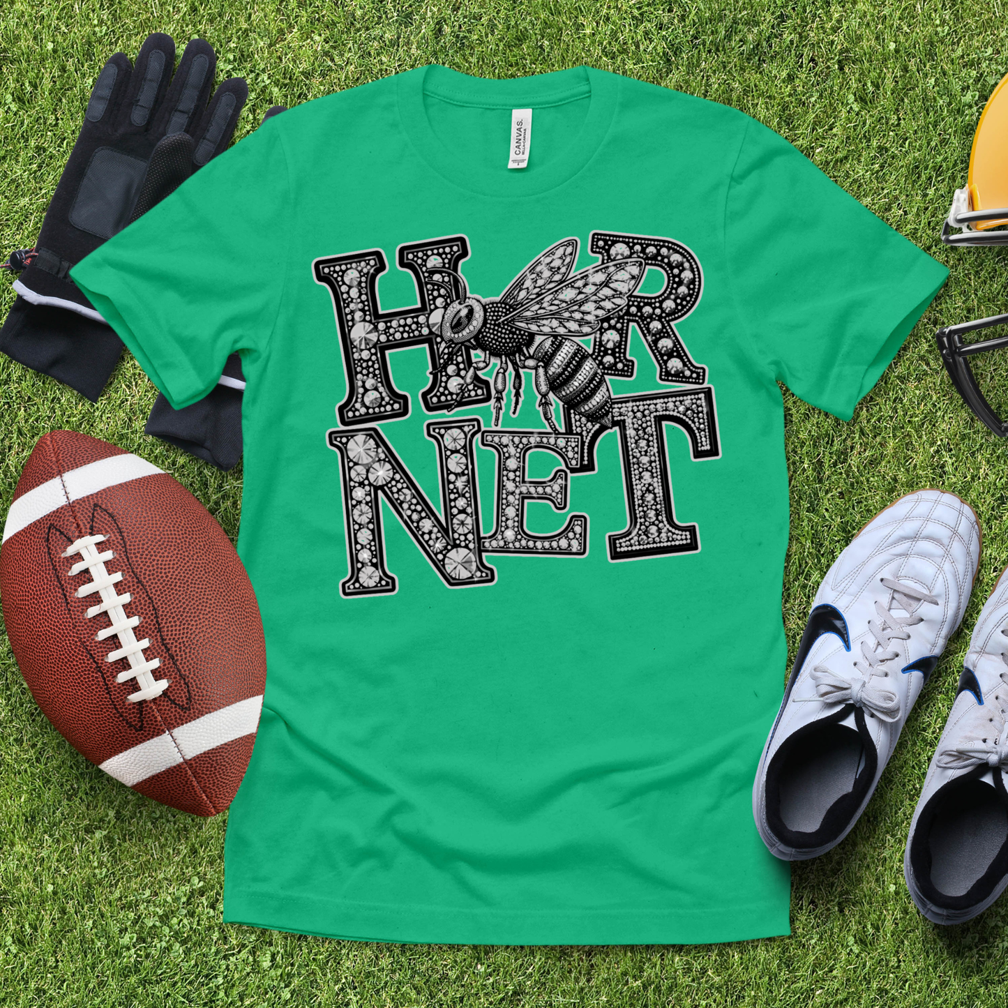 A green short-sleeve graphic T-shirt laid flat on artificial grass, surrounded by football, gloves, sneakers, and eyeglasses. The shirt has a large black-and-white 'HORN NET' text design with a hornet illustration across the chest.