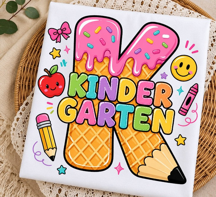 A square white fabric item, likely a napkin or small cloth square, printed with a colorful cartoon design featuring the word 'KINDER GARTEN' in pastel letters over two waffle cones shaped like pencils, surrounded by playful school-themed doodles. The cloth is styled on a woven wicker tray over a beige lace surface.