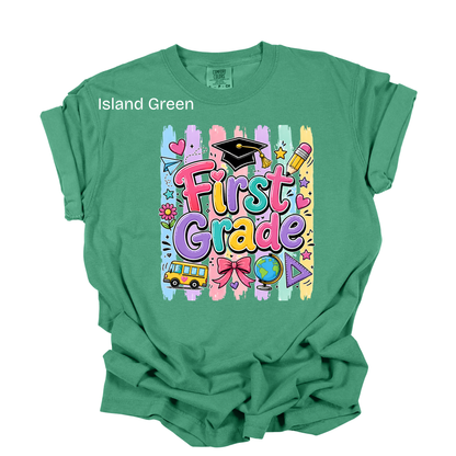 A green short-sleeve t-shirt with a colorful 'First Grade' graphic on the front, featuring a school bus, graduation cap, and paint-stroke background. The text 'Island Green' is printed on the upper left chest area.