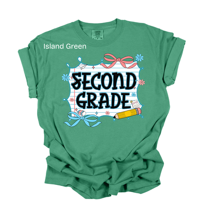A green short-sleeve t-shirt with a 'SECOND GRADE' graphic on the front, featuring school-themed illustrations like a pencil, ribbon, and chalkboard. The text 'Island Green' is printed near the left shoulder.