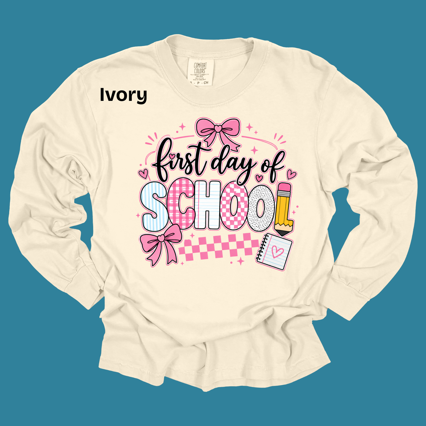 An ivory long-sleeve t-shirt with a 'first day of SCHOOL!' graphic on the front, featuring pink bows, checkered patterns, and school supplies, displayed against a solid teal background.