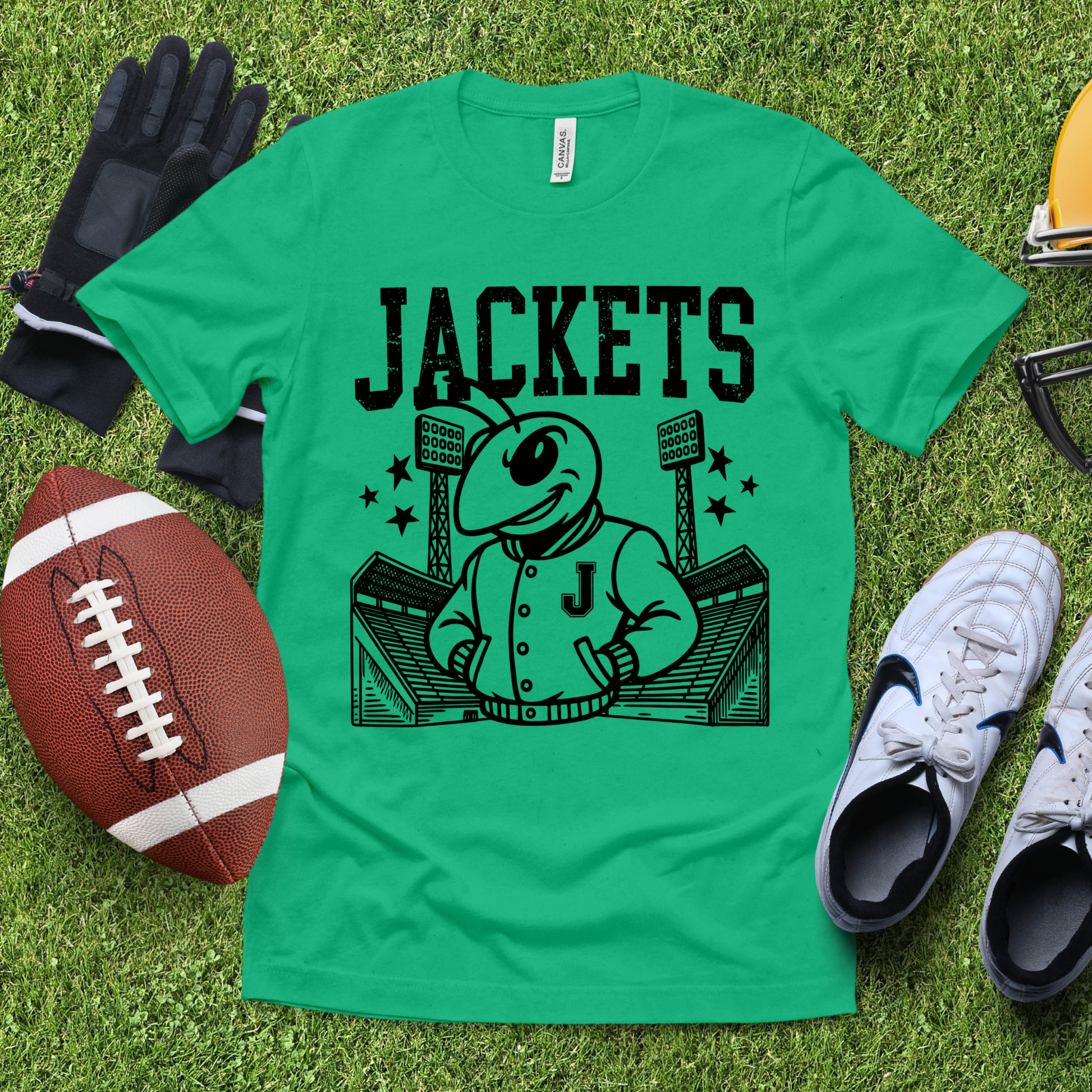 A bright green short-sleeve T-shirt laid flat on artificial grass, featuring a large black graphic of a cartoon bee mascot wearing a jacket with the letter J, under the word "JACKETS." Surrounding the shirt are football, gloves, sneakers, and a helmet as styled props.