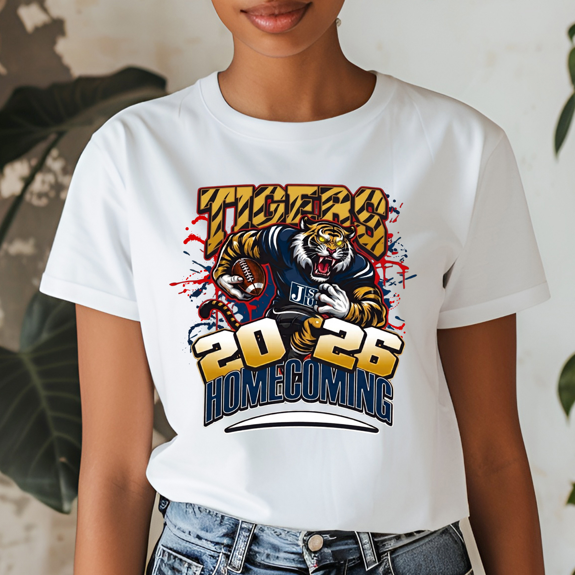 A woman wearing a white short-sleeve graphic T-shirt with a large tiger illustration and the text 'Tigers 2026 Homecoming' in yellow, blue, and red. The photo is cropped from the lower face to the hips, with jeans visible against a softly blurred indoor background with plants.