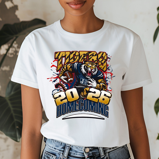 A woman wearing a white short-sleeve graphic T-shirt with a large tiger illustration and the text 'Tigers 2026 Homecoming' in yellow, blue, and red. The photo is cropped from the lower face to the hips, with jeans visible against a softly blurred indoor background with plants.
