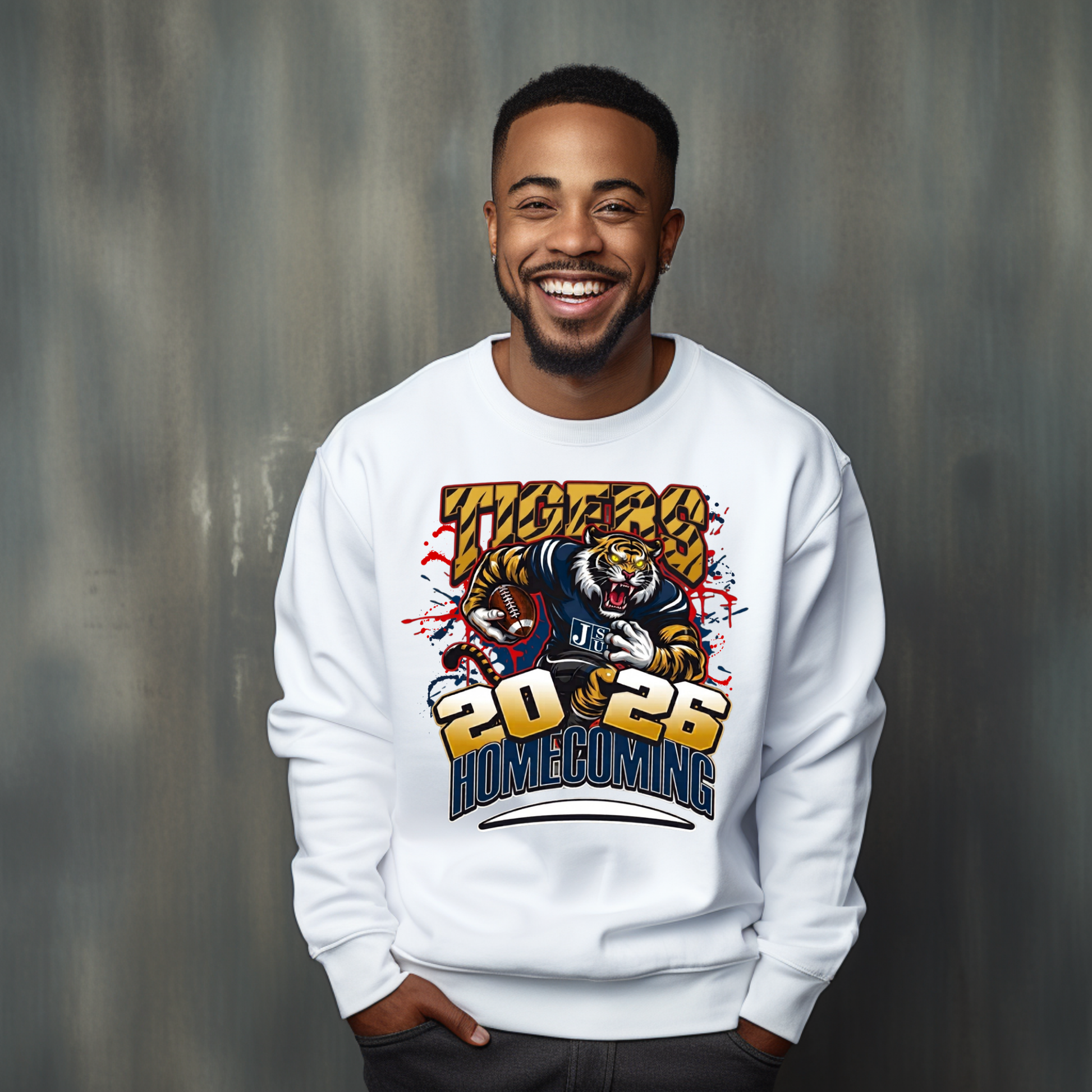 A smiling man wearing a white crewneck sweatshirt with a colorful graphic print featuring a tiger mascot and the text 'VICTORY 2026 HOMECOMING', posed against a textured gray studio background.